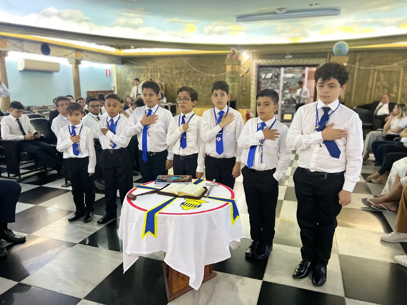 Escudeiros de camisa branca e gravata azul em cerimônia, com a mão ao peito ao redor de uma mesa