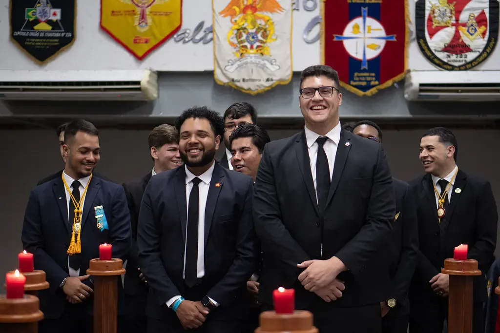 Sêniores DeMolay de terno reunidos diante dos estandartes, em cerimônia à luz de velas